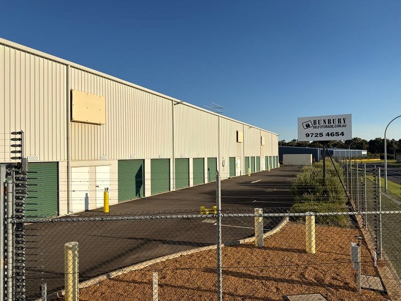 Row of green roller-door self storage units beside a paved driveway at an outdoor storage facility.