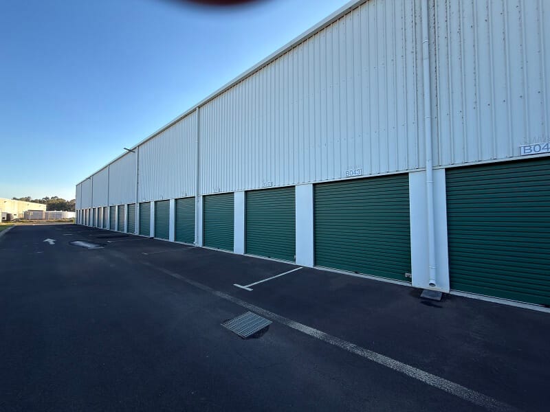 Row of green roller-door self storage units beside a paved driveway at an outdoor storage facility.