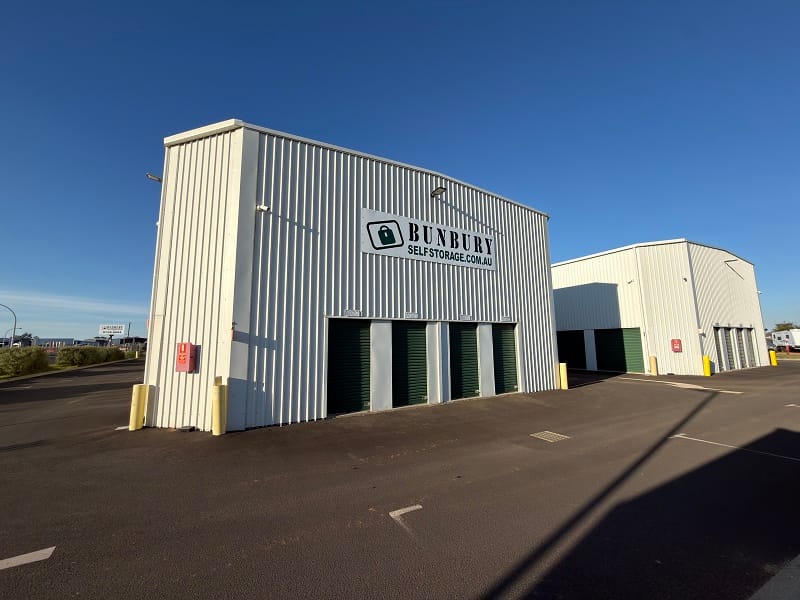 Row of green roller-door self storage units beside a paved driveway at an outdoor storage facility.