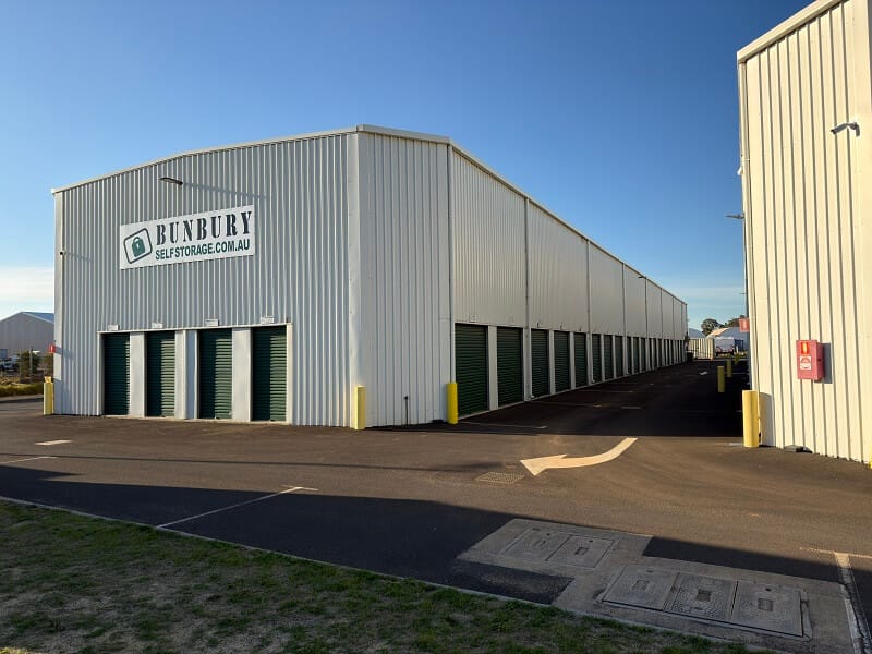 Row of green roller-door self storage units beside a paved driveway at an outdoor storage facility.