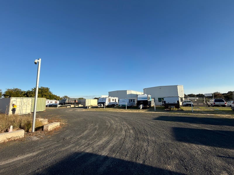 Row of green roller-door self storage units beside a paved driveway at an outdoor storage facility.